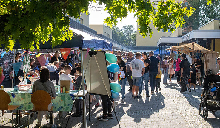 Ein lebhaftes Familien-Kiezfest im Freien in der Thermometersiedlung, bei dem sich Menschen aller Altersgruppen an Tischen, Ständen und Zelten bei strahlendem Sonnenschein versammeln. Dekorationen, Luftballons und schattige Plätze sorgen für eine festliche Atmosphäre auf dem gepflasterten Innenhof.