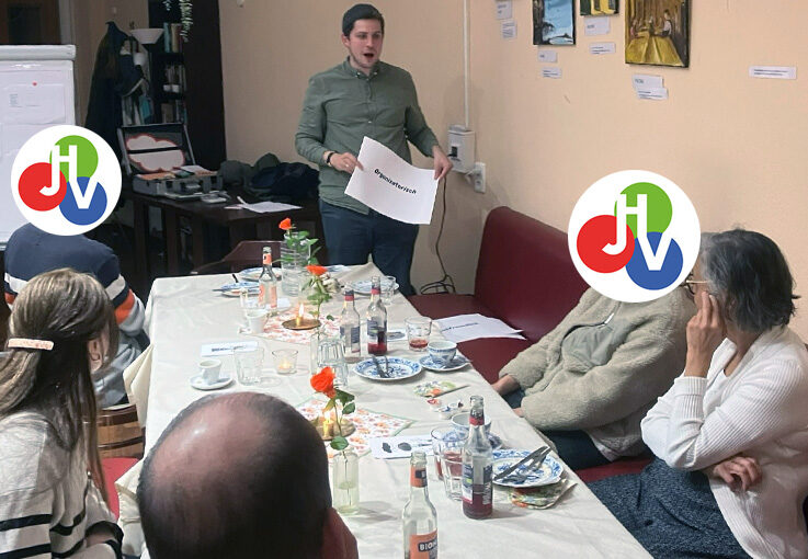 Ein Mann steht und präsentiert einer kleinen Gruppe, die um einen Tisch mit Getränken und Tellern in einer entspannten EinLaden-Einstellung sitzt. Einige Gesichter sind durch bunte Logos verdeckt, die dieser Szene voller Kunstwerke und einer Motivationsschub-Atmosphäre eine spielerische Note verleihen.