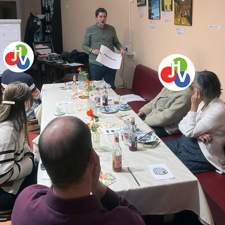 Ein Mann steht am Kopfende eines Tisches im einLaden, hält ein Blatt Papier in der Hand und gibt seinem Team einen Motivationsschub, während er vier sitzenden Personen, umgeben von Getränken, Tellern und Papieren, in einem gemütlichen Innenraum präsentiert.