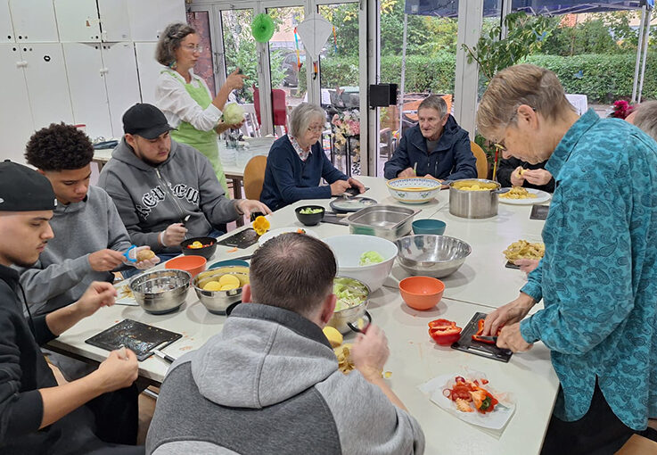Eine bunt gemischte Gruppe von Menschen, jung und alt, versammelt sich um einen Tisch und bereitet in einem hellen Raum mit großen Fenstern und viel Grün draußen gemeinsam Gemüse zu - eine Atmosphäre voller Wärme, ganz wie ein automatisch gespeicherter Entwurf von Zusammengehörigkeit.