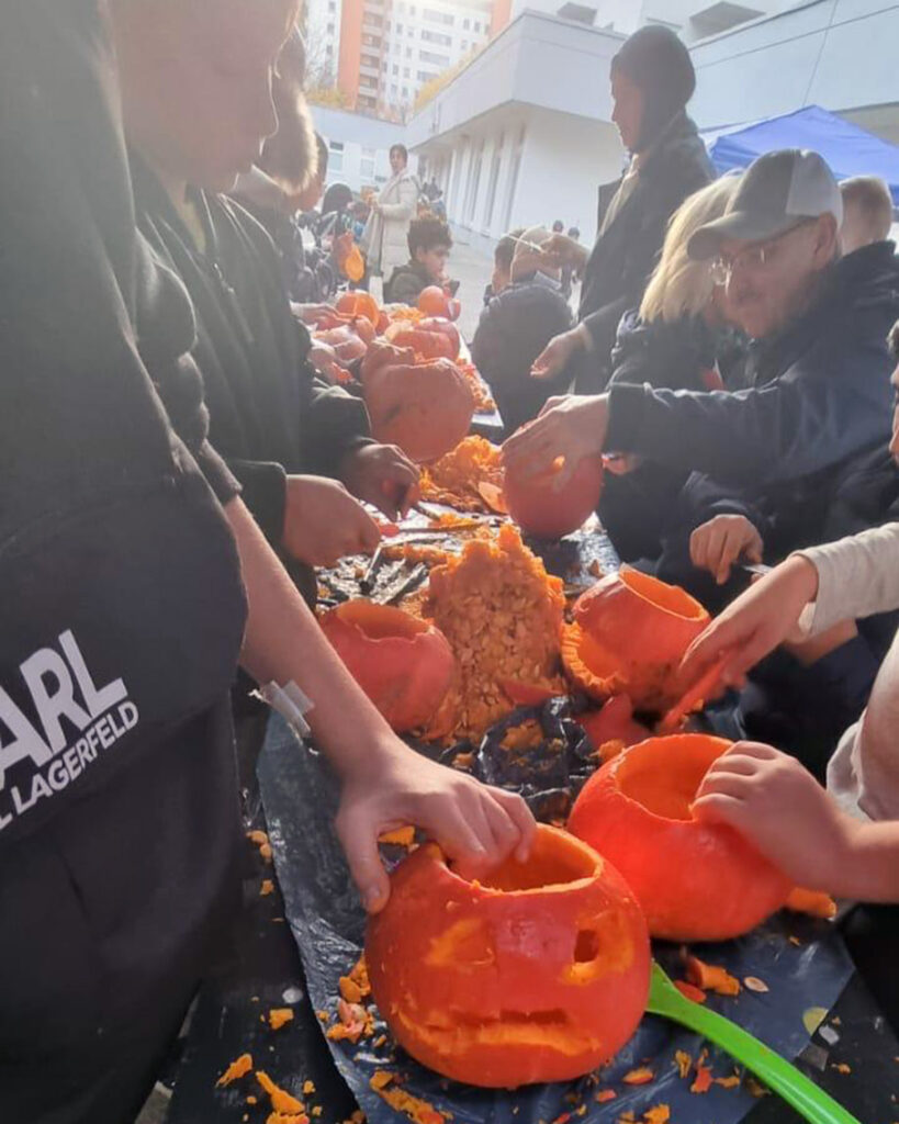Eine Gruppe von Menschen schnitzt Kürbisse an einem langen Tisch im Freien während einer lebhaften Halloween-Party im BUSSTOP Familienzentrum, umgeben von Kürbisstücken und Werkzeugen. Die geschnitzten Gesichter und das geschäftige Treiben lassen auf eine festliche Veranstaltung oder eine Werkstattatmosphäre schließen.