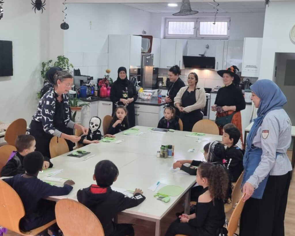 Kinder in schwarzen Kostümen sitzen bei der Halloween-Party des BUSSTOP Familienzentrums um einen Tisch und lauschen den verkleideten Erwachsenen. Spinnen und andere Dekorationen hängen von der Decke und schaffen eine lebendige und einnehmende Atmosphäre im Küchenklassenzimmer.