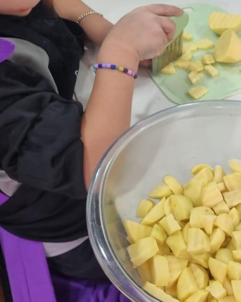 Im BUSSTOP Familienzentrum schneidet ein Kind mit einem Faltenschneider gelbes Gemüse auf einem Schneidebrett für die Halloween-Party. Das Kind trägt ein Perlenarmband und dunkle Kleidung mit einer lila Schürze, daneben steht eine Schüssel mit gewürfeltem Gemüse.