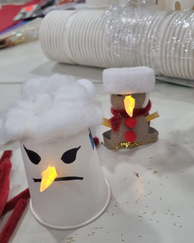 Zwei handgefertigte Schneemänner aus dem BUSSTOP Familienzentrum: einer aus einer umgedrehten weißen Tasse mit Baumwolle oben drauf, schwarzem Marker-Gesicht und leuchtender gelber Nase; der andere aus Filz mit Baumwollhut und leuchtender Nase. Perfekt für Ihre Halloween-Party!.