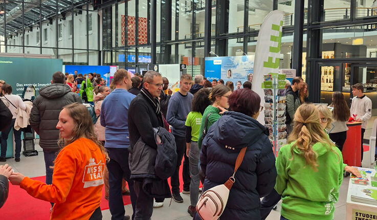 Eine geschäftige Indoor-Veranstaltung auf der ABI-Zukunft-Berlin in einem modernen Glasgebäude, mit Menschen, die sich unterhalten und Messestände besuchen. Einige Teilnehmer tragen grüne und orangefarbene Hemden mit Logos, und überall sind Informationsdisplays und Banner zu sehen.