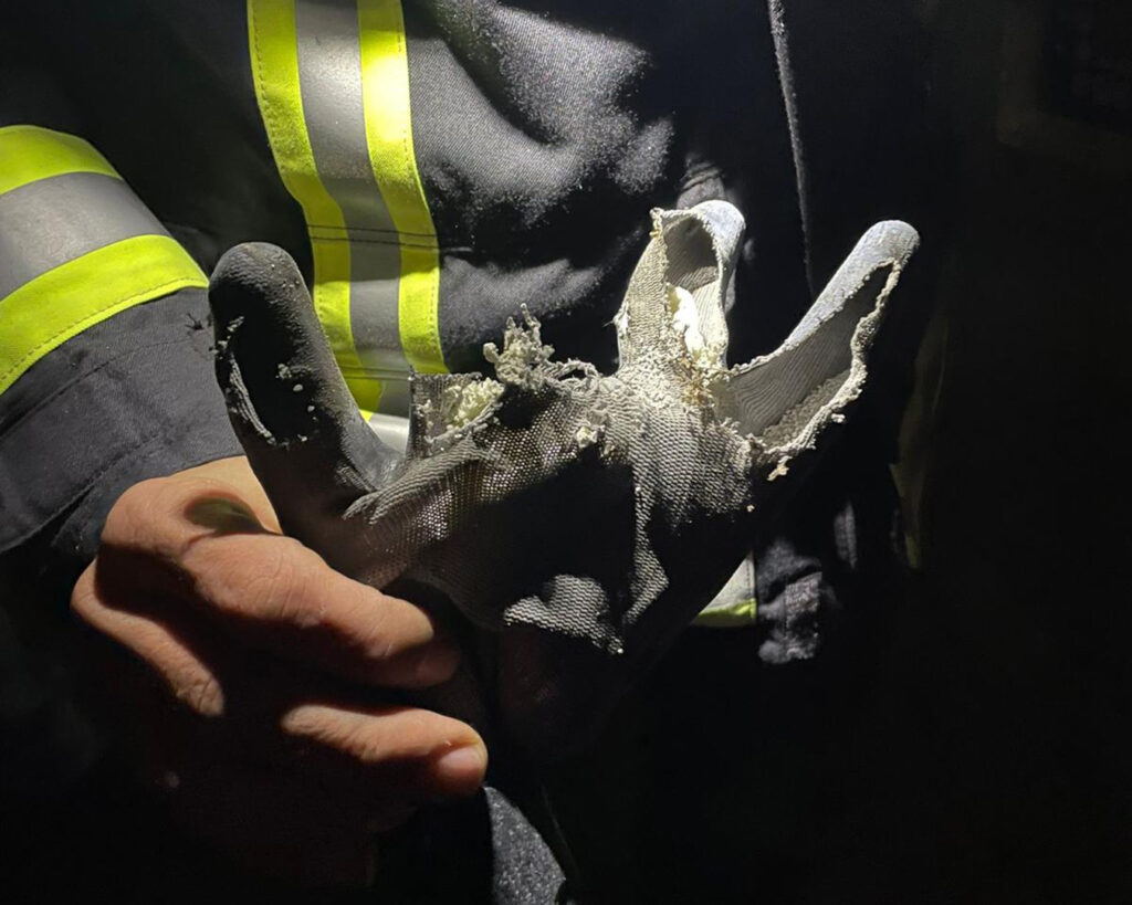 Eine Person in einer dunklen Jacke mit reflektierenden Streifen hält einen stark beschädigten, teilweise geschmolzenen Handschuh unter einen Lichtstrahl - ein automatisch gespeicherter Entwurf der starken Abnutzungs- und Brandspuren des Handschuhs wird hervorgehoben.