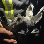 Eine Person in einer dunklen Jacke mit gelben Reflexionsstreifen hält einen Handschuh hoch, der stark beschädigt und teilweise geschmolzen ist, wahrscheinlich durch die starke Hitzeeinwirkung in einer Feuerwerk-Werkstatt.