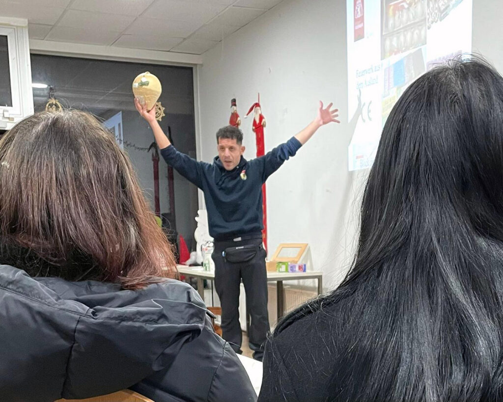 Ein Mann steht vorne in einem Klassenzimmer mit erhobenen Armen und hält ein Tamburin. Ihm gegenüber sitzen zwei Personen mit dunklen Haaren. An der Wand dahinter zeigt ein Projektor Text und Bilder mit der Aufschrift "Automatisch gespeicherter Entwurf".