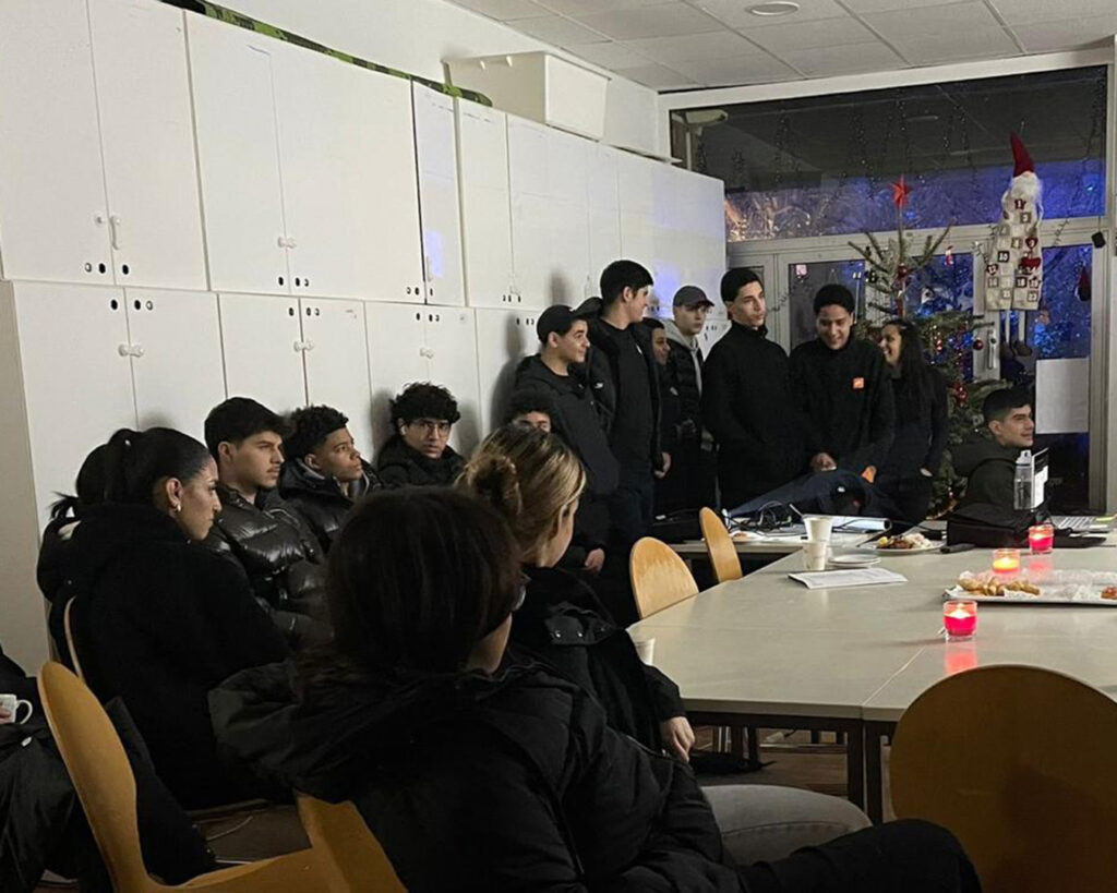 Eine Gruppe junger Leute, meist schwarz gekleidet, versammelt sich um Tische in einem Raum, der mit einem Weihnachtsbaum und einer Weihnachtsmannfigur geschmückt ist. Papiere und brennende Kerzen deuten auf einen automatisch gespeicherten Entwurf oder ein Treffen hin, das stattfindet.