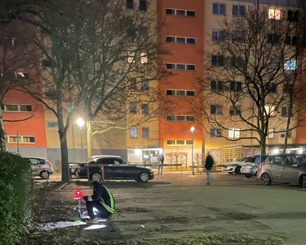 Eine Person in einer reflektierenden Weste hockt nachts auf einer schwach beleuchteten Straße und leuchtet mit einer Taschenlampe auf den Boden - ein automatisch gespeicherter Entwurf, bei dem im Hintergrund Gebäude, Autos, Bäume und ein paar Menschen zu sehen sind.