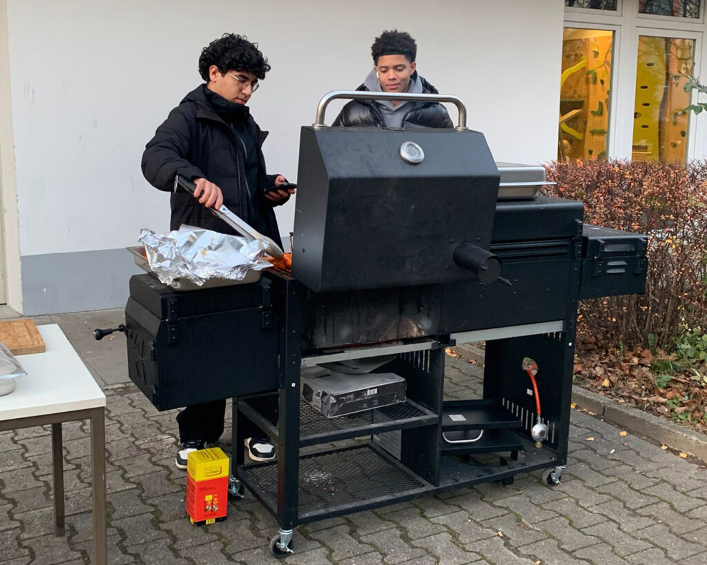 Zwei Menschen stehen draußen an einem großen schwarzen Grill; einer kocht mit einer Zange, während der andere zusieht, als ob er einen automatisch gespeicherten Entwurf begutachtet. Die Szene spielt sich in der Nähe eines Gebäudes mit Büschen und Herbstlaub im Hintergrund ab.