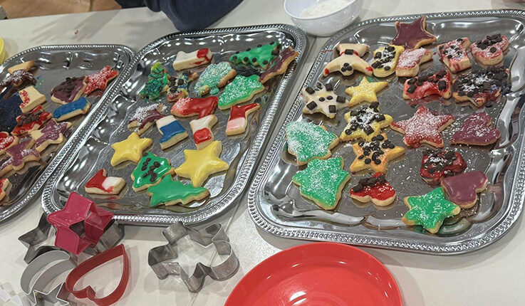 Drei Tabletts mit dekorierten Weihnachtsplätzchen - Sterne, Bäume und Tiere - schmücken den Tisch mit buntem Zuckerguss und Streuseln. Ein roter Teller und sternförmige Ausstechformen schaffen eine festliche Szene, perfekt für ein lebendiges Adventskalendertreffen.
