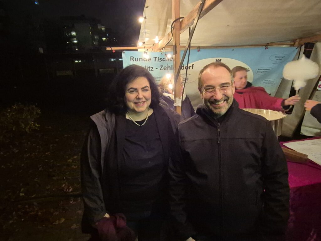 Zwei Personen stehen lächelnd nebeneinander auf einem nächtlichen Wintermarkt 2025, unter warmen Lichterketten in der Nähe eines Standes. Im Hintergrund sind andere Personen und ein Schild mit deutschem Text zu sehen.