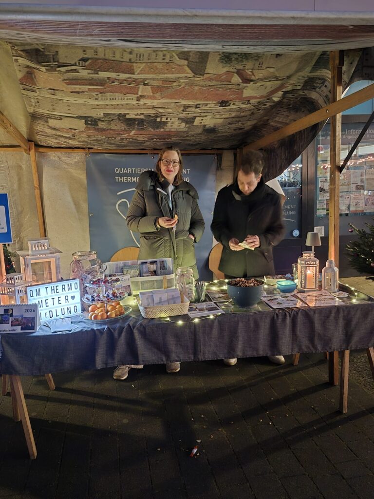 Zwei Personen stehen hinter einem festlichen Stand auf dem Wintermarkt 2025, der mit Lichterketten beleuchtet und mit Snacks und Dekorationsartikeln gefüllt ist. Unter einem Vordach ist auf dem Tisch vor ihnen ein Schild mit der Aufschrift Thermometer zu sehen.