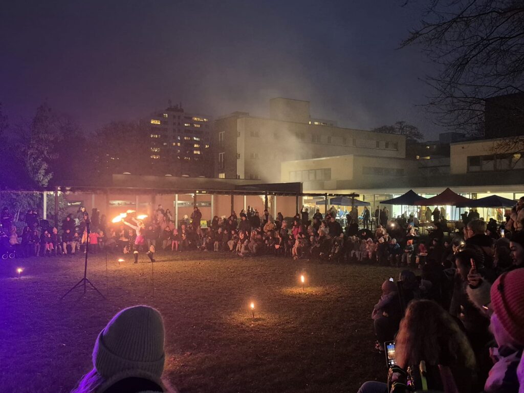 Ein Feuerkünstler unterhält eine große Menschenmenge, die während des Wintermarkts 2025 nachts im Freien sitzt, mit Gebäuden und Zelten im Hintergrund. Das Publikum schaut gebannt zu, während kleine Feuer auf dem Gras den Auftrittsbereich markieren.