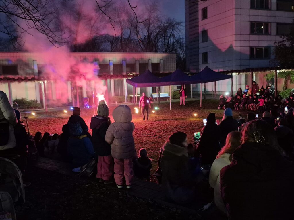 Eine Menschenmenge sitzt in der Abenddämmerung während des Wintermarkts 2025 im Freien und beobachtet einen Darsteller vor einem Gebäude. Bunte Lichter und Rauch erzeugen einen dramatischen Effekt. Einige Kinder stehen vorne, während Erwachsene in Winterkleidung auf dem Boden sitzen oder knien.