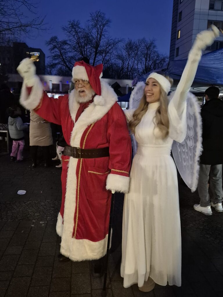 Der Weihnachtsmann und eine als Engel verkleidete Frau stehen während des Wintermarkts 2025 nachts im Freien, lächeln und winken. Im Hintergrund sind Menschen und ein Zelt zu sehen, außerdem kahle Bäume und ein Gebäude.