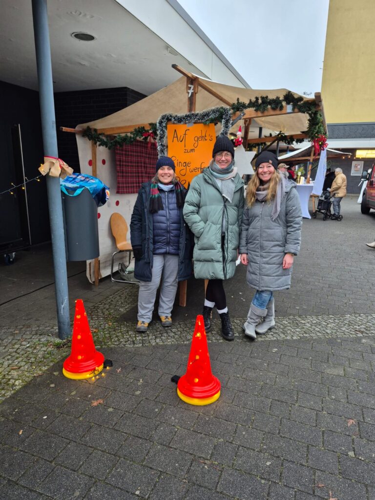 Drei Personen in Wintermänteln und -mützen stehen lächelnd vor einem dekorierten Holzstand mit einem gelben Wintermarkt 2025-Schild. Auf dem Boden davor stehen zwei orangefarbene Sicherheitskegel, im Hintergrund sind weitere Stände und Menschen zu sehen.