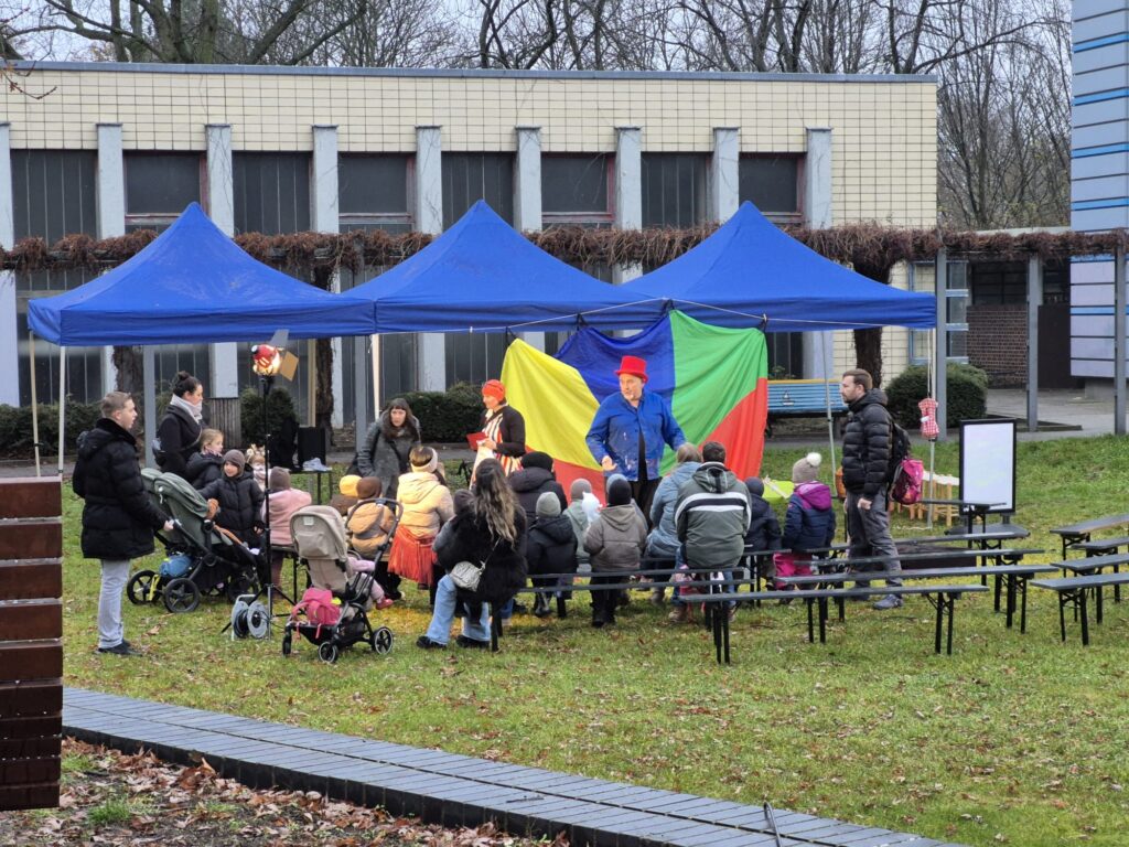 Eine Gruppe von Kindern und Erwachsenen sitzt auf Bänken im Freien unter blauen Zelten auf dem Wintermarkt 2025 und beobachtet zwei Künstler vor einer farbenfrohen Kulisse. Die Veranstaltung findet auf einer Wiese mit einem modernen Gebäude im Hintergrund statt.