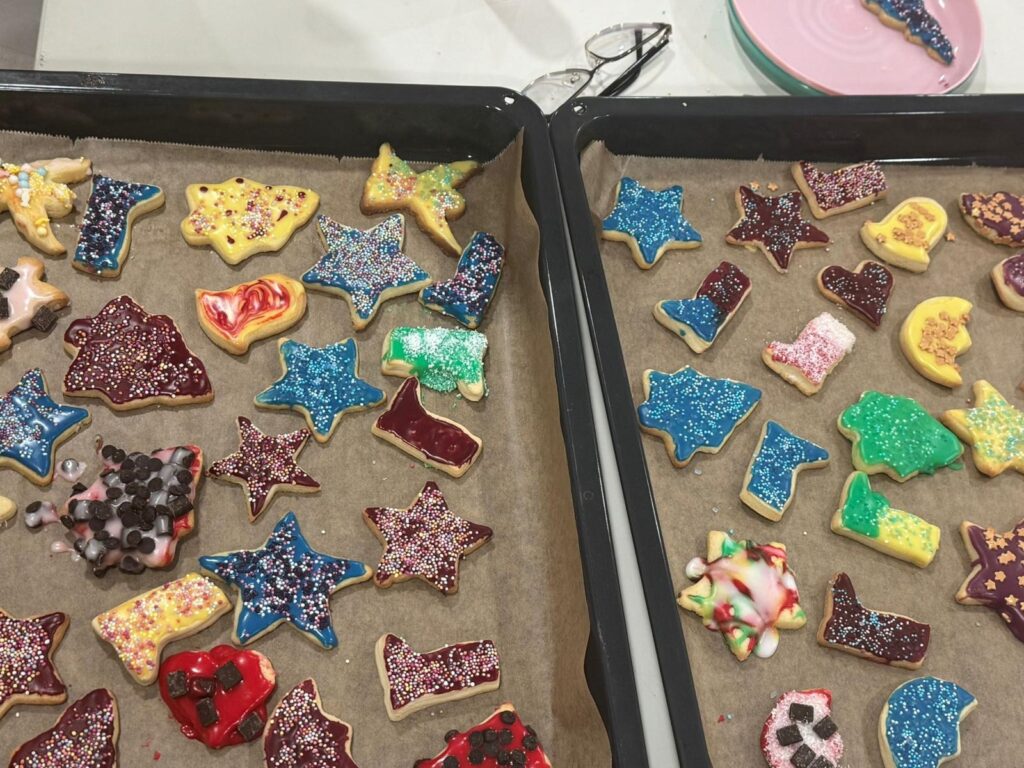 Zwei Backbleche, gefüllt mit verschiedenen, bunt verzierten Zuckerplätzchen - ein automatisch gespeicherter Entwurf von Sternen, Stiefeln, Tieren und Herzen -, die mit buntem Zuckerguss und Streuseln überzogen sind. Ein paar Gläser und ein rosa Teller stehen daneben.
