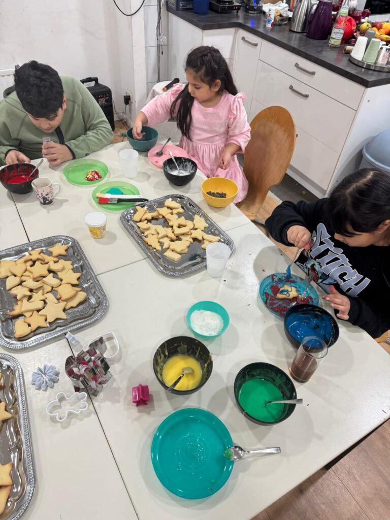 Drei Kinder sitzen an einem Tisch und verzieren Weihnachtsplätzchen mit buntem Zuckerguss und Streuseln. In der hellen Küche sind verschiedene Schüsseln mit Zuckerguss, Ausstechformen, Tabletts mit unverzierten Plätzchen und ein automatisch gespeicherter Entwurf ausgebreitet.