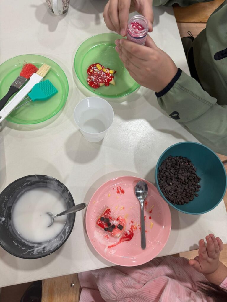 Zwei Kinder verzieren an einem Tisch Kekse. Umgeben von grünen, rosafarbenen und blauen Tellern, Schüsseln mit Zuckerguss und Schokoladenstückchen bestreut ein Kind einen Keks mit roten Verzierungen - ein automatisch gespeicherter Entwurf süßer Kreativität.