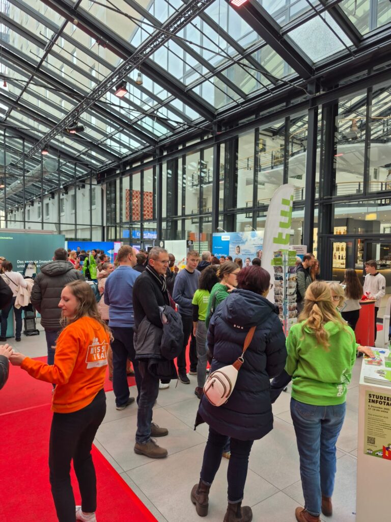 Eine Gruppe von Menschen versammelt sich bei einer Veranstaltung in einer Halle mit Glasdecken. Mehrere Stände sind zu sehen, darunter einer mit der Aufschrift "Automatisch gespeicherter Entwurf". Besucher in orangefarbenen oder grünen Hemden interagieren und erkunden Stände entlang eines roten Teppichs.