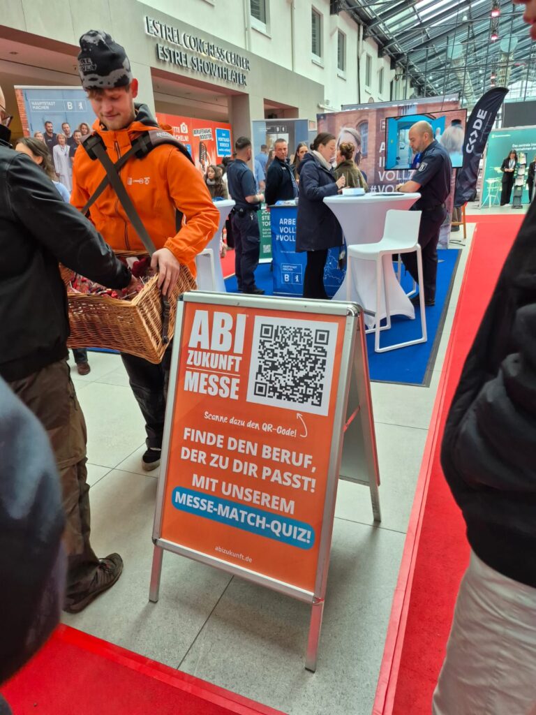 Eine geschäftige Karrieremesse mit Menschen, die an Ständen interagieren. Im Vordergrund wirbt ein Schild für die ABI Zukunft Messe mit einem QR-Code und der Aufforderung, durch ein Quiz einen passenden Beruf zu finden. Automatisch gespeicherter Entwurf wird neben einem Mann mit einem Korb angezeigt.