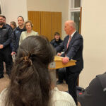 Ein Mann im Anzug spricht auf einem Podium im Abgeordnetenhaus zu einem Raum voller Menschen, einige in Uniform. Ein Kameramann filmt von rechts, während die Gäste dieser Dankeschön-Einladung aufmerksam zuhören.