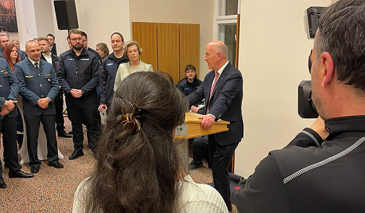 Ein Mann im Anzug spricht auf einem Podium im Abgeordnetenhaus zu einem Raum voller Menschen, einige in Uniform. Ein Kameramann filmt von rechts, während die Gäste dieser Dankeschön-Einladung aufmerksam zuhören.