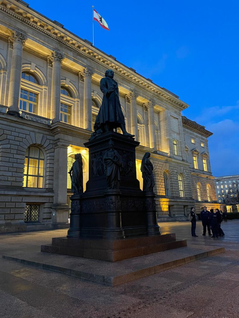 Eine von kleineren Figuren umgebene Bronzestatue steht in der Abenddämmerung vor einem beleuchteten neoklassizistischen Gebäude, auf dessen Dach eine Fahne weht - eine Szene, die an einen automatisch gespeicherten Entwurf erinnert, der zum Leben erweckt wird, während sich einige Menschen in der Nähe versammeln.