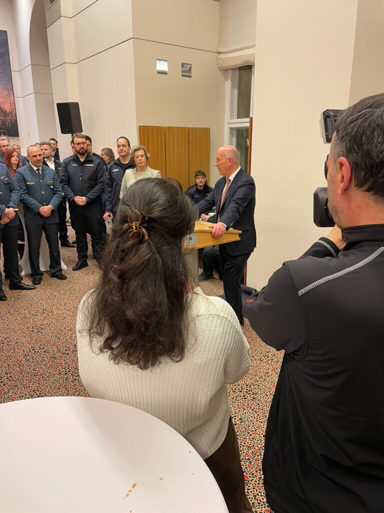 Ein Mann im Anzug spricht von einem Podium aus zu einer Gruppe von Menschen, darunter auch Polizisten, die sich in einem Raum versammelt haben. Während ein Fotograf diesen automatisch gespeicherten Entwurf festhält, hören einige der Anwesenden an einem kleinen Tisch aufmerksam zu.