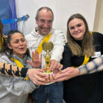 Fünf lächelnde Teammitglieder stehen dicht beieinander und halten jeweils eine kleine goldene Trophäe. Sie tragen gelbe Bänder und scheinen in einer zwanglosen Umgebung einen Mannschaftssieg zu feiern.