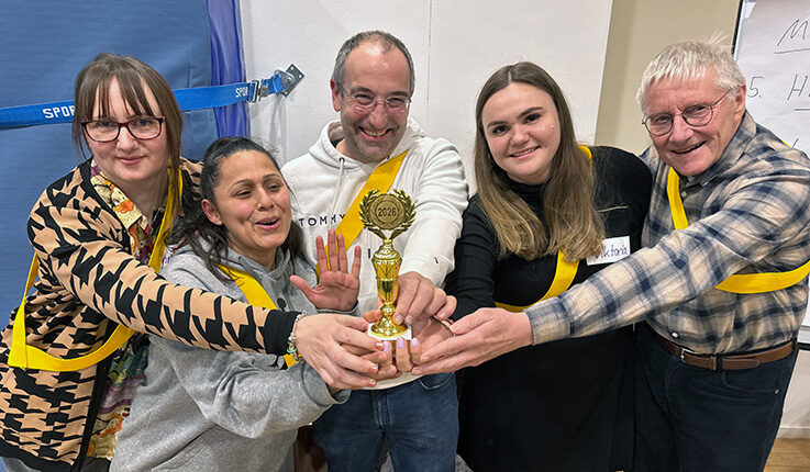 Fünf lächelnde Teammitglieder stehen dicht beieinander und halten jeweils eine kleine goldene Trophäe. Sie tragen gelbe Bänder und scheinen in einer zwanglosen Umgebung einen Mannschaftssieg zu feiern.