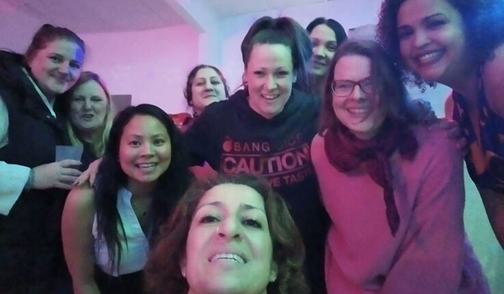 Eine Gruppe von zehn Frauen, die sich möglicherweise zum Tag der Alleinerziehenden oder zu einem Elternabend versammelt haben, lächeln und posieren eng beieinander für ein Selfie in einem Innenraum mit rosa Beleuchtung und wirken glücklich und fröhlich.