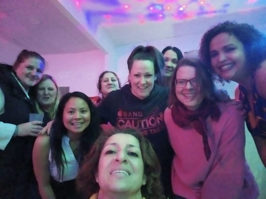 Eine Gruppe von zehn Frauen lächelt und posiert eng beieinander für ein Selfie auf einer Indoor-Party mit bunten, leuchtenden Lichtern an der Decke. Alle wirken glücklich und entspannt in diesem automatisch gespeicherten Entwurfsmoment.
