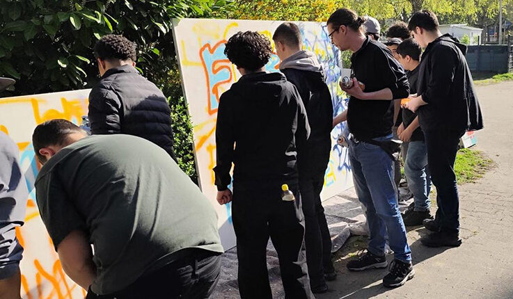 Bei einem lebhaften Graffiti-Workshop steht eine Gruppe von Menschen im Freien vor großen weißen Tafeln und sprüht bunte Graffiti. Die sonnige Szene im Grünen fängt die Konzentration und den Erfolg der Teilnehmer auf ihr Kunstwerk ein.