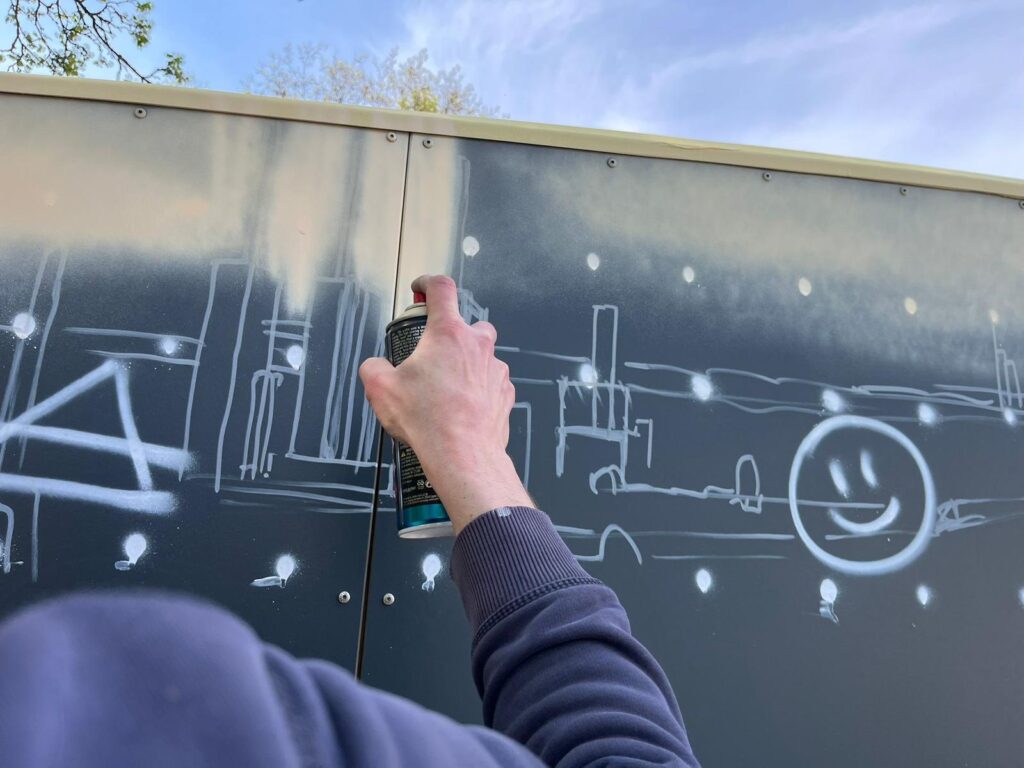Die Hand einer Person, die eine Sprühdose hält und eine weiße Stadtlandschaft und ein Smiley-Gesicht während eines Graffiti-Workshops auf einer dunklen Fläche im Freien unter einem teilweise bewölkten Himmel kreiert, fängt urbane Kreativität und Erfolg ein.