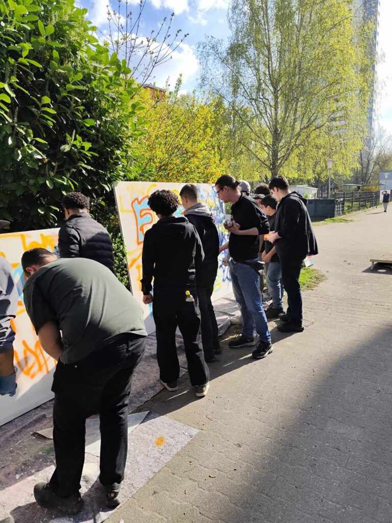 Eine Gruppe von Menschen steht bei einem Graffiti-Workshop im Freien und malt an einem sonnigen Tag Graffiti auf große weiße Tafeln, die entlang eines von grünen Büschen und Bäumen gesäumten Bürgersteigs aufgestellt sind - ein voller Erfolg!.