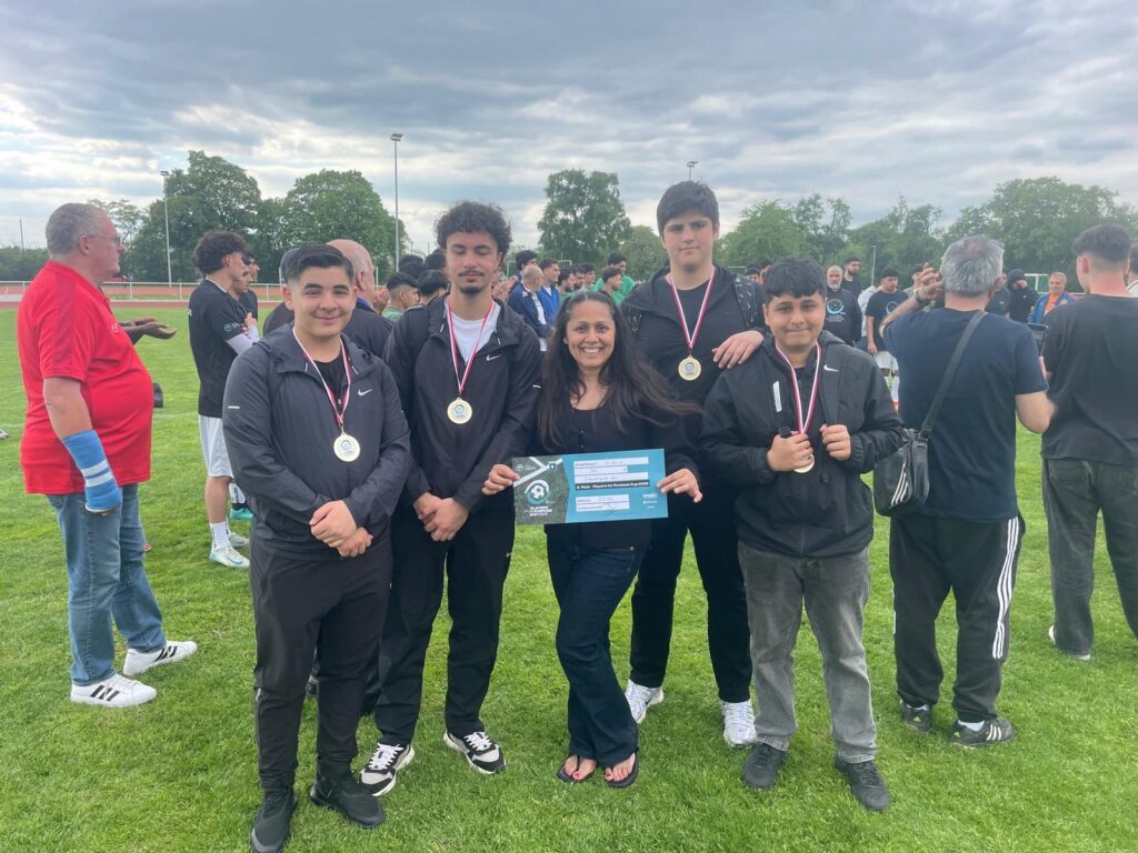 Fünf Personen stehen zusammen auf einer Wiese - vier junge Männer mit Medaillen und in schwarzer Kleidung und eine Frau mit einer Urkunde für den Players for Purpose Cup. Stolz feiern sie ihren 6. Platz und das Preisgeld von 750 Euro unter einem wolkenverhangenen Himmel.