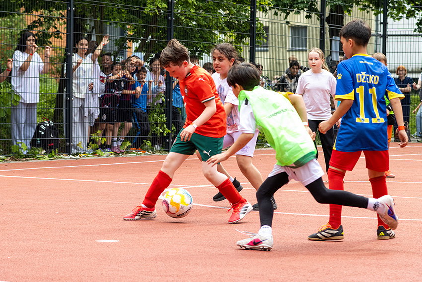 Kinder spielen ein Fußballspiel auf einem Platz im Freien, einige in roten Shorts, andere in grünen oder blauen. Die Zuschauer jubeln hinter einem Zaun und fotografieren das Geschehen. Üppiges Grün umgibt diese lebhafte Szene - schnell wie ein automatisch gespeicherter Entwurf.