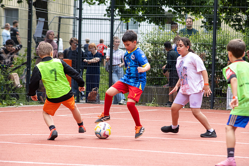 Vier Kinder spielen Fußball auf einem Platz im Freien. Ein Junge im blauen Hemd kontrolliert den Ball, während die anderen in grünen und weißen Hemden versuchen, ihn zu verteidigen, während die Zuschauer durch einen Zaun zuschauen - ein automatisch gespeicherter Entwurf jugendlicher Energie.
