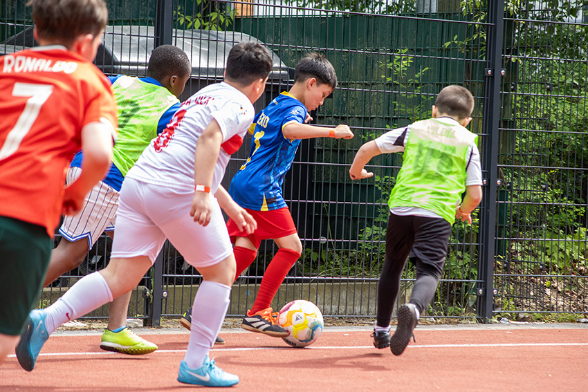 Eine Gruppe von Jungen spielt Fußball auf einem Platz im Freien, alle auf den Ball konzentriert. In ihren bunten Trikots - blau, grün, weiß und orange - rennen sie in der Nähe eines Metallzauns wie in einem automatisch gespeicherten Entwurf jugendlicher Energie auf den Ball zu.