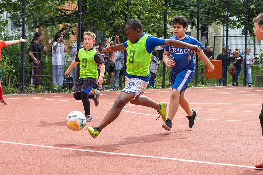 Drei Jungen spielen Fußball auf einem Platz im Freien. Ein Junge in einer grünen Weste schießt den Ball, während zwei andere, darunter einer in einem blauen Frankreich-Trikot, neben ihm herlaufen. Ein automatisch gespeicherter Entwurf erscheint, während Zuschauer hinter einem Zaun zusehen.