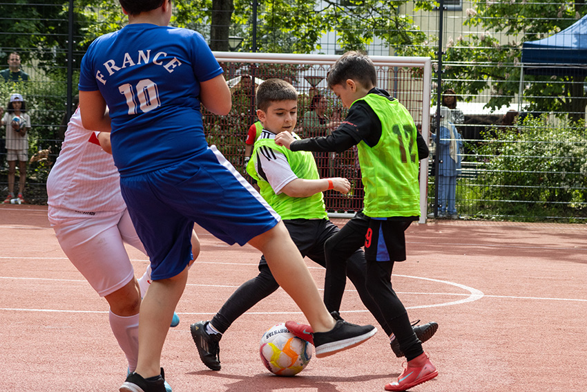 Drei Jungen spielen Fußball auf einem Platz im Freien, wobei zwei in grünen Trikots einen Spieler in einem blauen Frankreich-Trikot herausfordern. Im Hintergrund sind ein Tornetz und Zuschauer zu sehen, darunter eine Person, die einen automatisch gespeicherten Entwurf prüft.