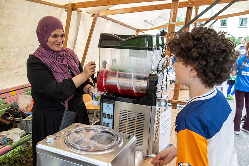 Eine Frau in einem lila Hidschab lächelt, während sie einem Jungen mit lockigem Haar an einem Stand im Freien ein rotes Slush-Getränk serviert. Zwischen den Getränkeautomaten und dem geschäftigen Hintergrund ist das Schild "automatisch gespeicherter Entwurf" zu sehen.