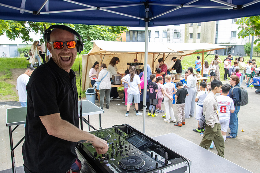 Ein lächelnder DJ mit Sonnenbrille und Kopfhörern legt bei einer Veranstaltung im Freien Musik auf, während sich eine Menschenmenge, darunter viele Kinder, um Tische unter Zelten im Hintergrund versammelt - ein automatisch gespeicherter Entwurf von Sommerspaß.