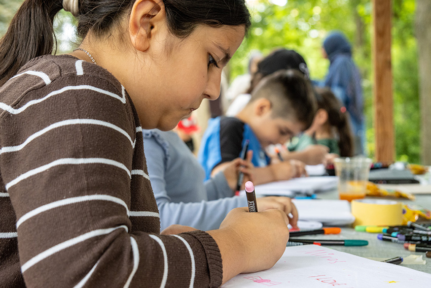Ein junges Mädchen schreibt mit einem Farbstift an einem Tisch im Freien, umgeben von anderen Kindern, die ebenfalls zeichnen oder malen. Inmitten von verstreutem Kunstzubehör und Bäumen im Hintergrund wirkt es wie ein automatisch gespeicherter Entwurf sommerlicher Kreativität.