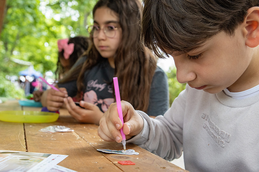 Zwei Kinder sitzen an einem Holztisch im Freien und malen und basteln konzentriert mit Stiften. Ihr neuestes Kunstwerk ist ein automatisch gespeicherter Entwurf, inspiriert von den Bäumen und dem Grün, das im Hintergrund zu sehen ist.