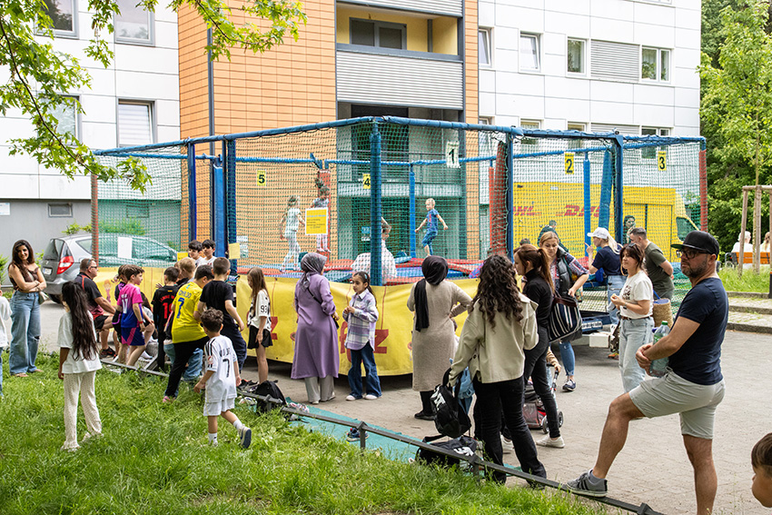Eine Gruppe von Kindern und Erwachsenen versammelt sich draußen in der Nähe eines Trampolinspielplatzes, in dem mehrere Kinder springen. Vor einem Wohnhaus stehen Menschen, die sich unterhalten und zuschauen - ein automatisch gespeicherter Entwurf eines lebendigen Gemeinschaftsvergnügens.